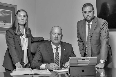 Office scene with a seated man signing documents at a desk, flanked by a woman and a man in suits.